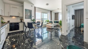 Un apartamento moderno con cocina, comedor y sala de estar. Cuenta con piso de mármol negro, muebles de cocina blancos y un amplio ventanal que proporciona luz natural.