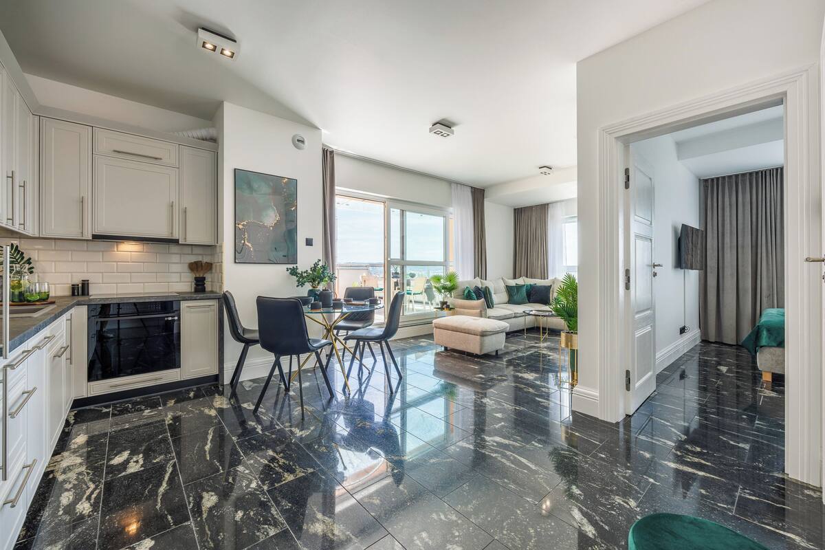 A modern apartment with a kitchen, dining area, and living room. It features a black marble floor, white cabinetry, and a large window providing natural light.