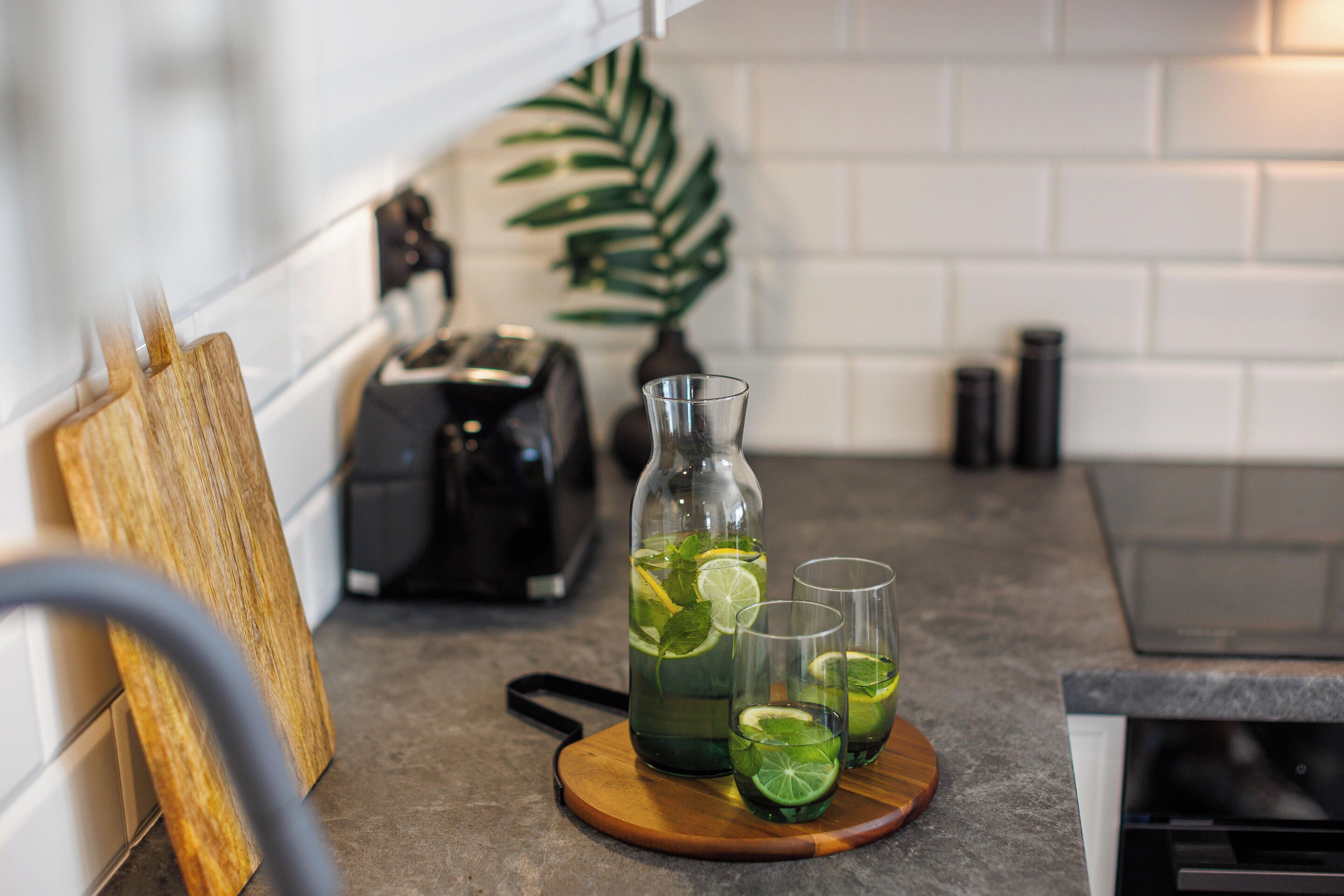 A kitchen counter with a wooden cutting board, a glass pitcher, and two glasses containing a green beverage with lime slices.