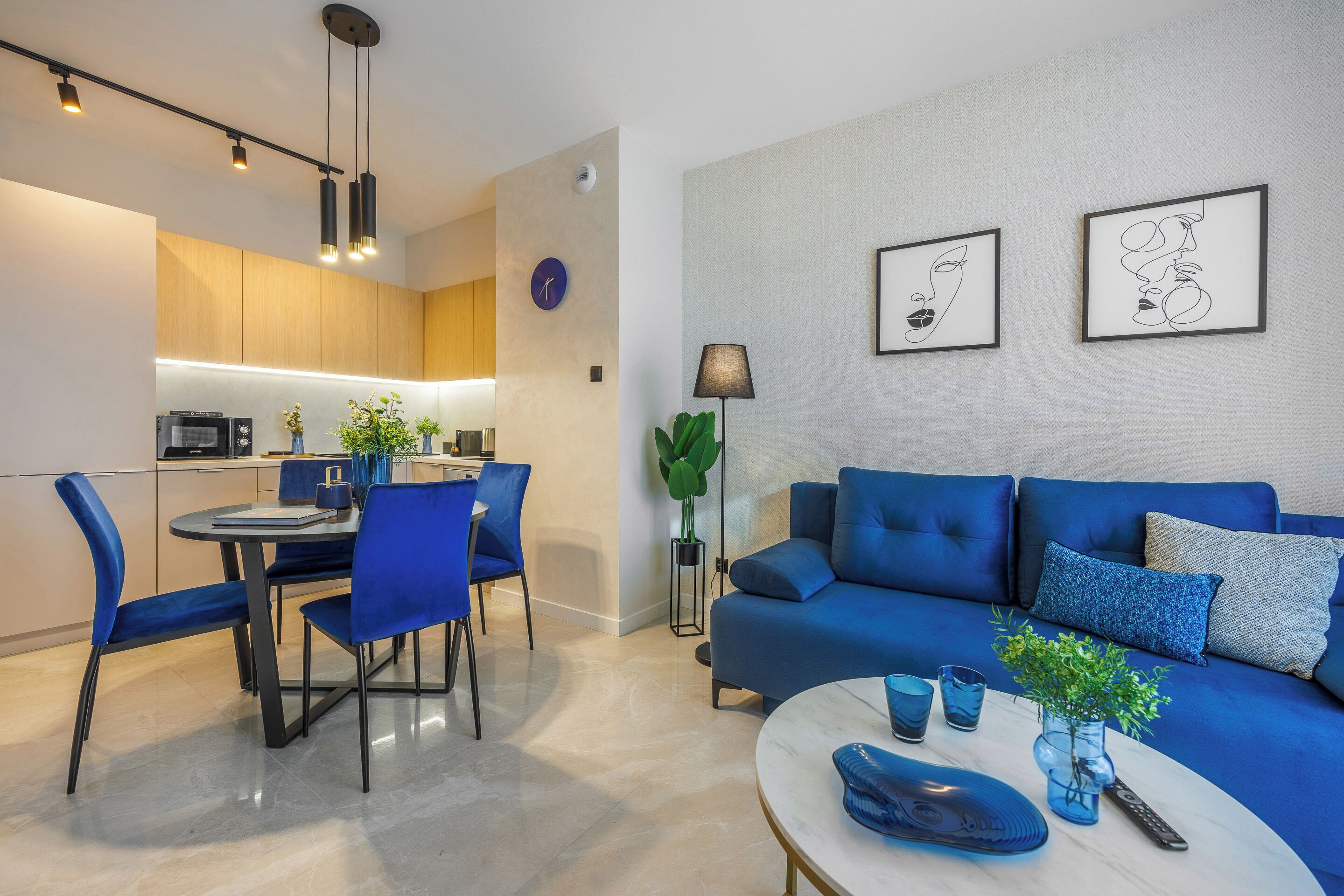 A modern living room with a blue sofa, a dining area with blue chairs, and a kitchen with wooden cabinets and a built-in oven.