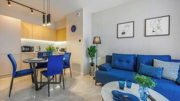 A modern living room with a blue sofa, a dining area with blue chairs, and a kitchen with wooden cabinets and a built-in oven.