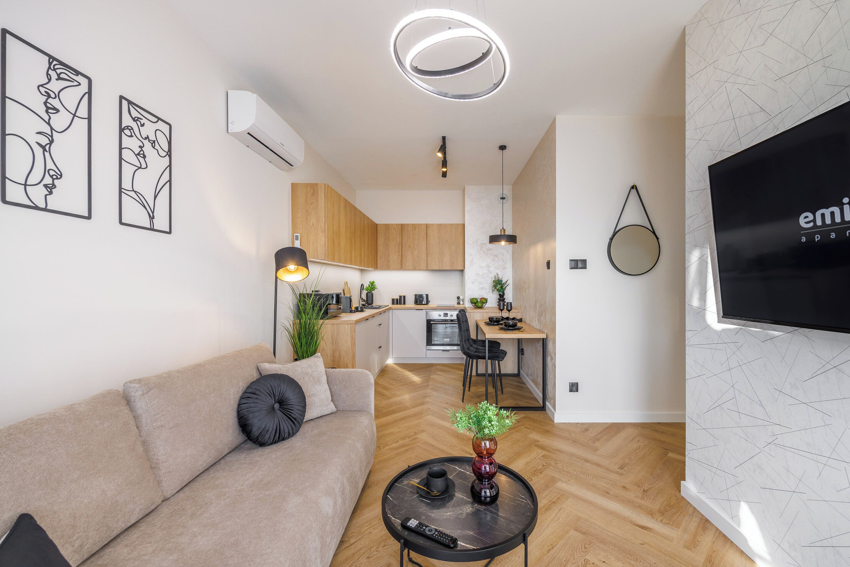 Una sala de estar moderna con un sofá, una mesa de centro con un florero y un control remoto, una cocina al fondo y un televisor montado en la pared.