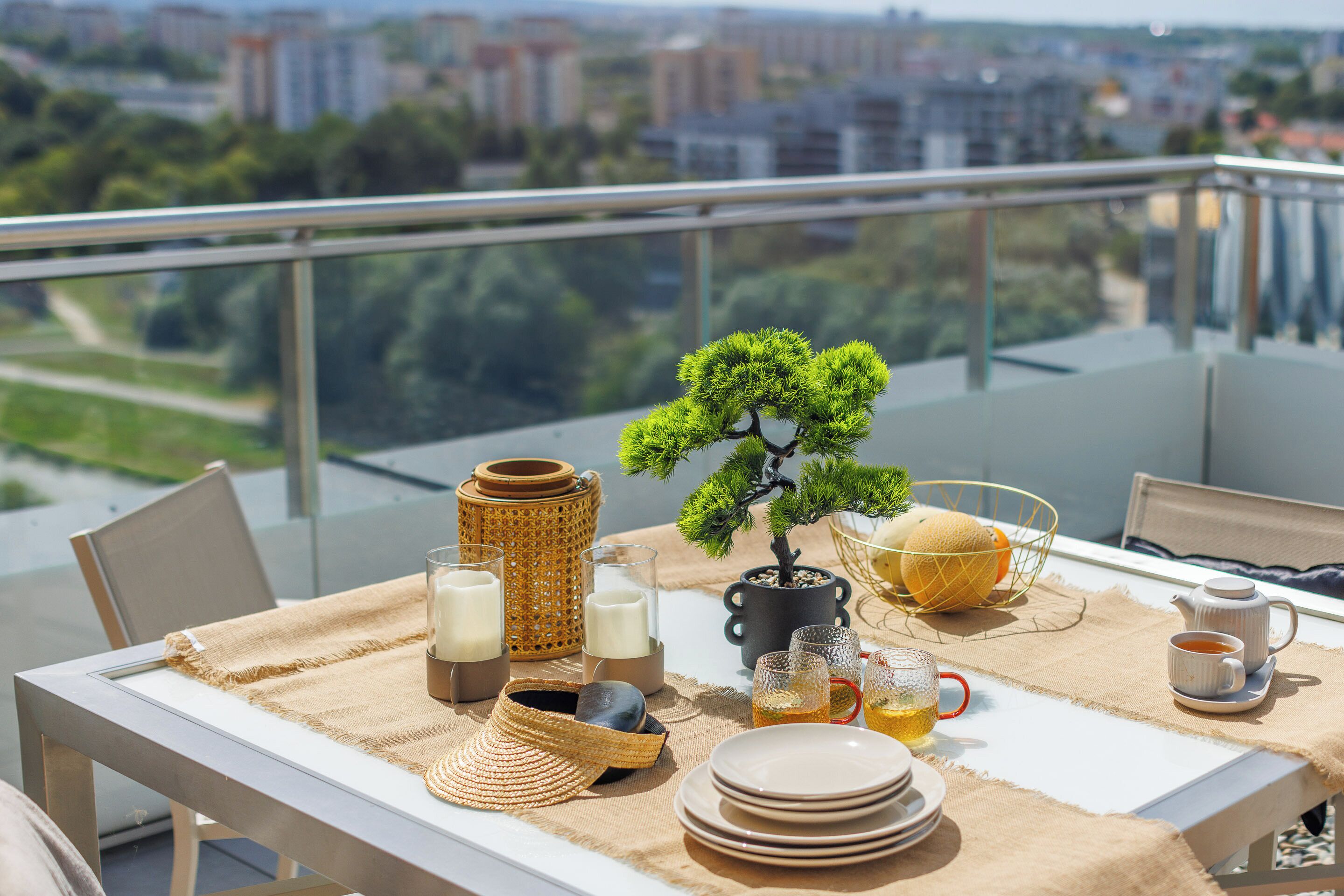 A balcony with a table set for tea, featuring a bonsai tree, a woven basket, and a view of the city.