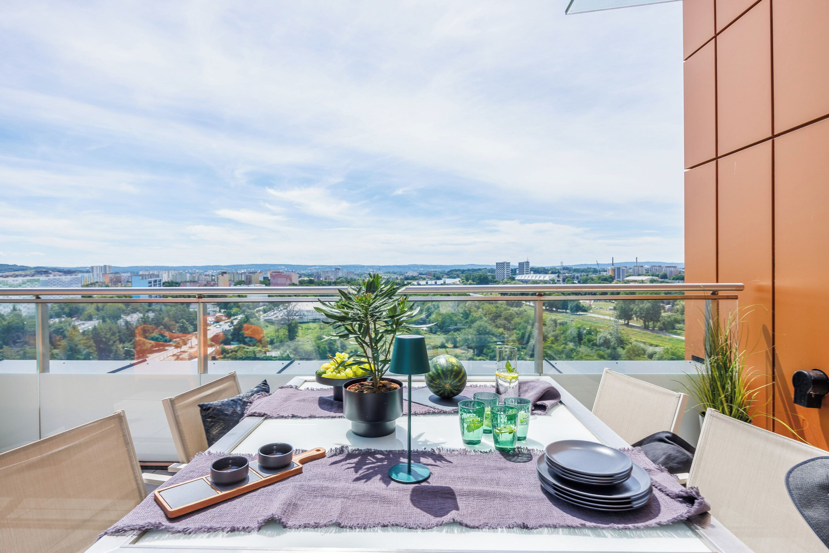 Uma varanda com mesa posta para refeições, um vaso de planta e vista para a cidade.