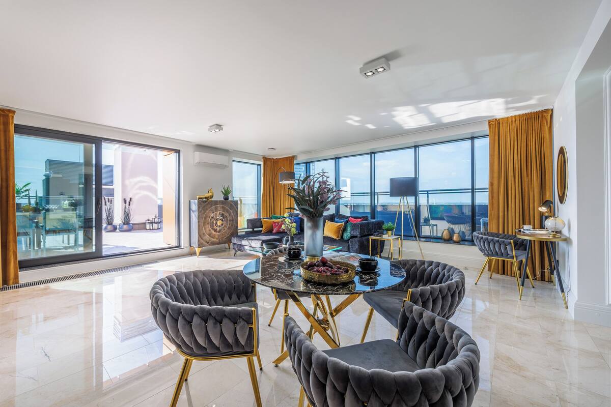 A spacious living room with a modern design, featuring a round table, plush chairs, and large windows offering a view of the city.