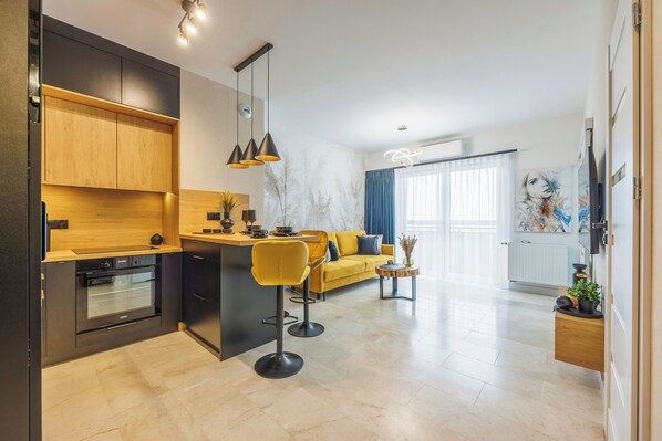 A modern kitchen with a built-in oven, a bar area with stools, a yellow sofa, and a large window with curtains. - Emihouse Riverside Apartments (Rzeszów)