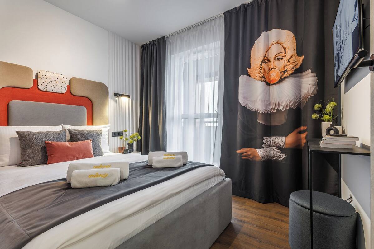 A modern hotel room with a large bed, a headboard with a unique design, a large window with sheer curtains, and a wall-mounted TV.