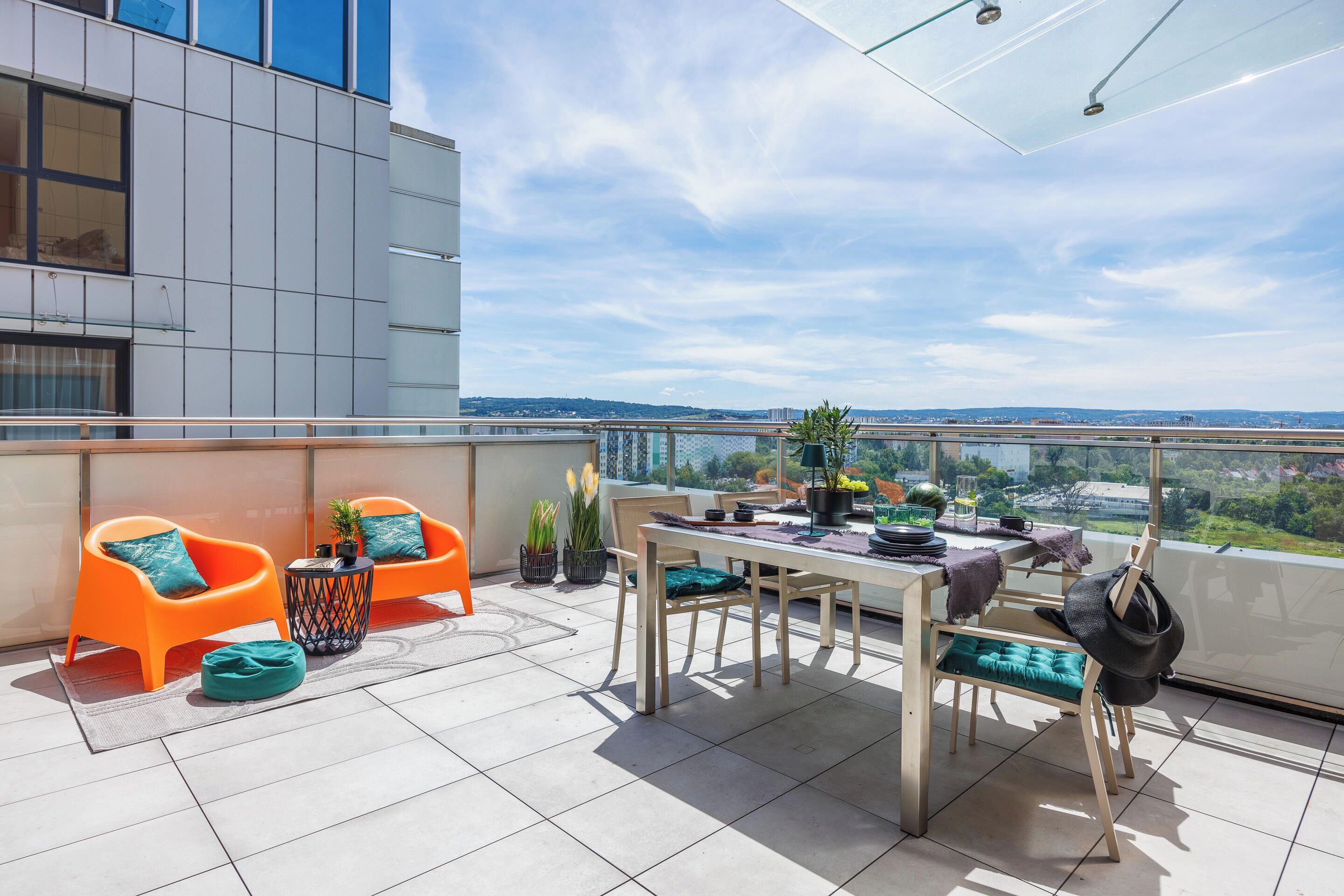 Una terraza moderna al aire libre con una mesa de comedor, dos sillas color naranja, una mesita negra y una planta en maceta.