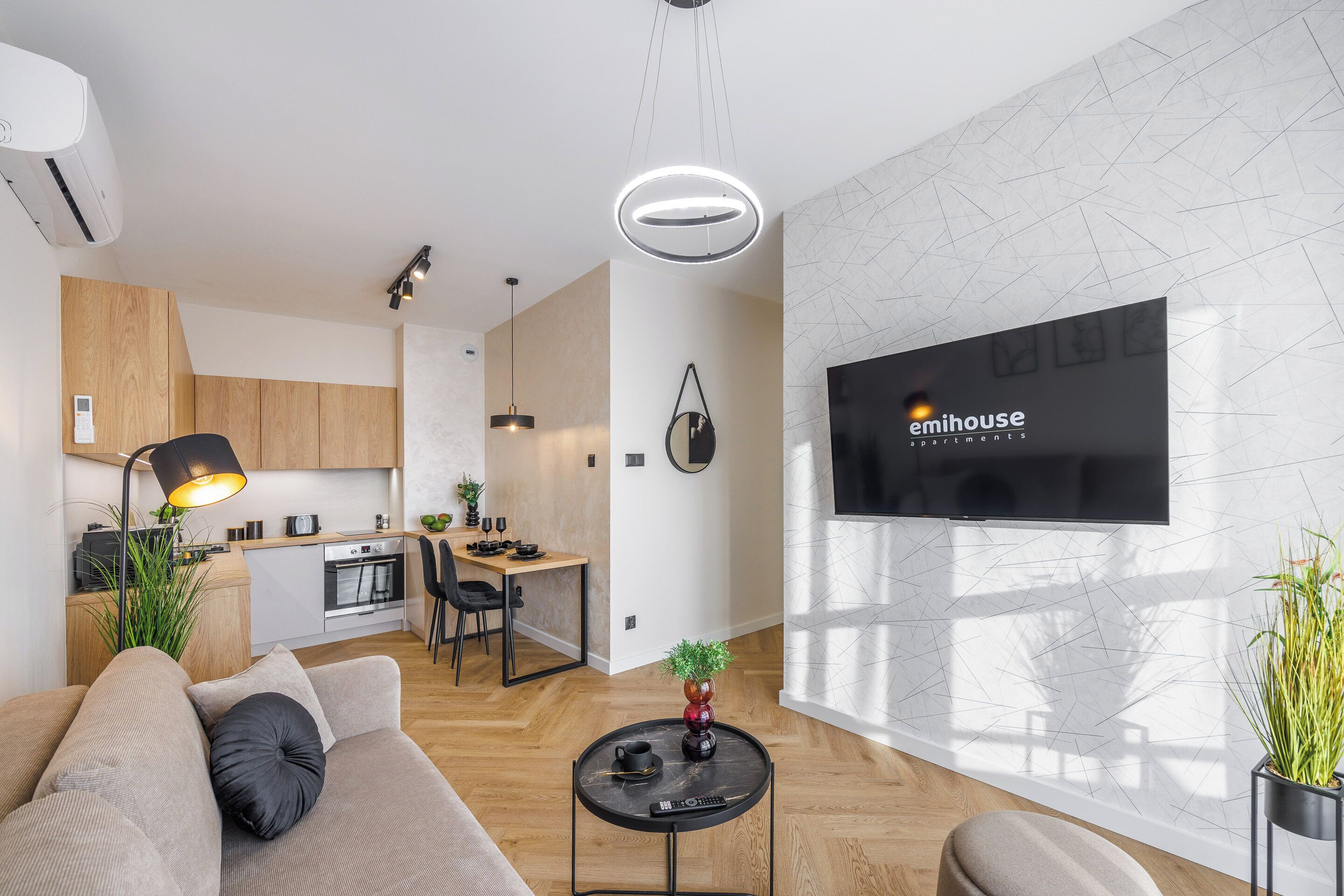 A modern apartment with a kitchen, dining area, and living room. A flat-screen TV is mounted on the wall, and there are plants and a coffee table in the living area.