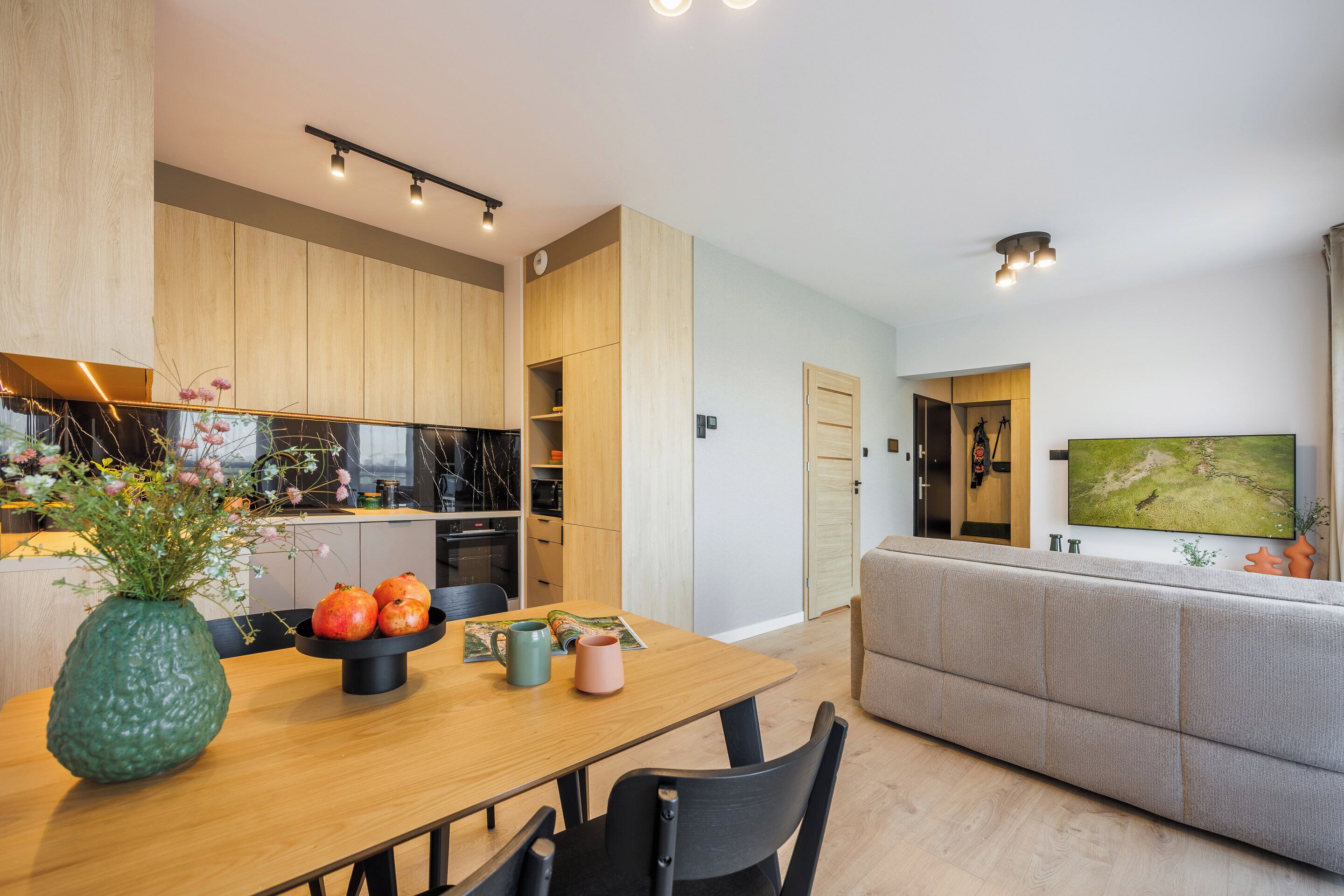 A modern kitchen with wooden cabinets, a dining table with chairs, and a living area with a sofa and a TV.