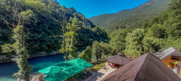 A view of a mountainous area with a clear blue river, surrounded by dense forest. There are wooden structures with green canopies and a small pavilion. - Rafting Camp Encijan (Foca)