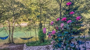 A hammock tied between trees by a calm lake, surrounded by lush greenery and blooming pink flowers. - Rafting Camp Encijan (Foca)