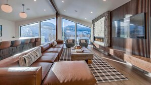 Un salon moderne avec un grand canapé, deux fauteuils, une table basse et une cheminée. De grandes fenêtres offrent une vue sur les montagnes.