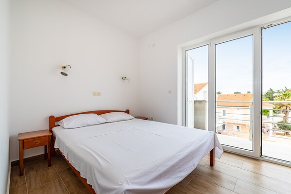 A bedroom with a bed, a nightstand, a window with a view of buildings, and a door. - Room-cozy Room Near Beach D (Novalja)