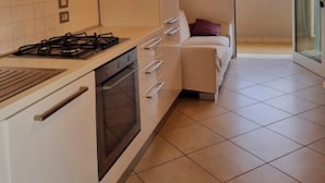Een moderne keuken met witte kasten, een gasfornuis en een inbouwoven. Er is een schuifdeur die leidt naar een balkon met uitzicht op de lucht en gebouwen.
