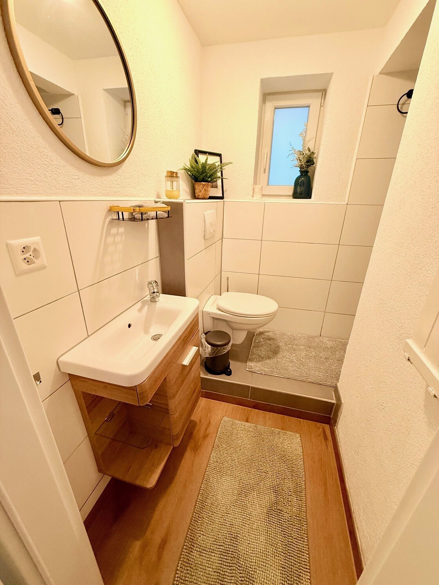 Ett badrum med ett vitt handfat, en toalett och en dusch. Det finns en spegel på väggen, ett litet fönster och en krukväxt på fönsterbrädan.