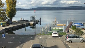 A parking area with cars and a dock by a calm lake, a flag on a small building, and mountains in the distance.