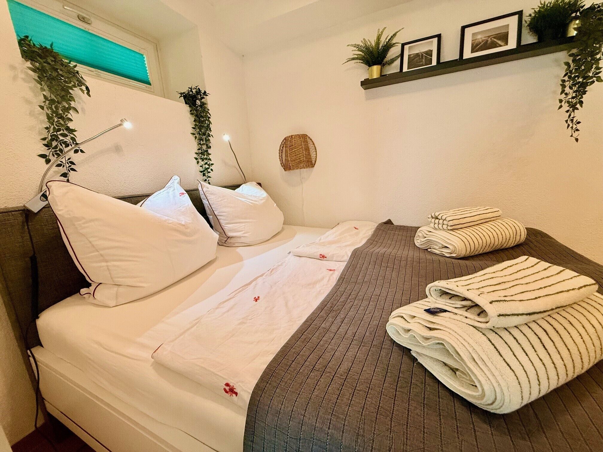 Un lit bien fait, avec du linge de lit blanc et une serviette à rayures, une lampe de chevet et une étagère ornée de plantes et de photos.