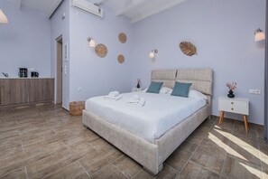 A bedroom with a bed, bedside tables, wall decor, and a kitchenette area. - GIN Wellness Hotel (Santorini)