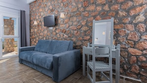 A modern room with a stone wall, a grey sofa, a small table with a mirror, and a TV mounted on the wall. - GIN Wellness Hotel (Santorini)