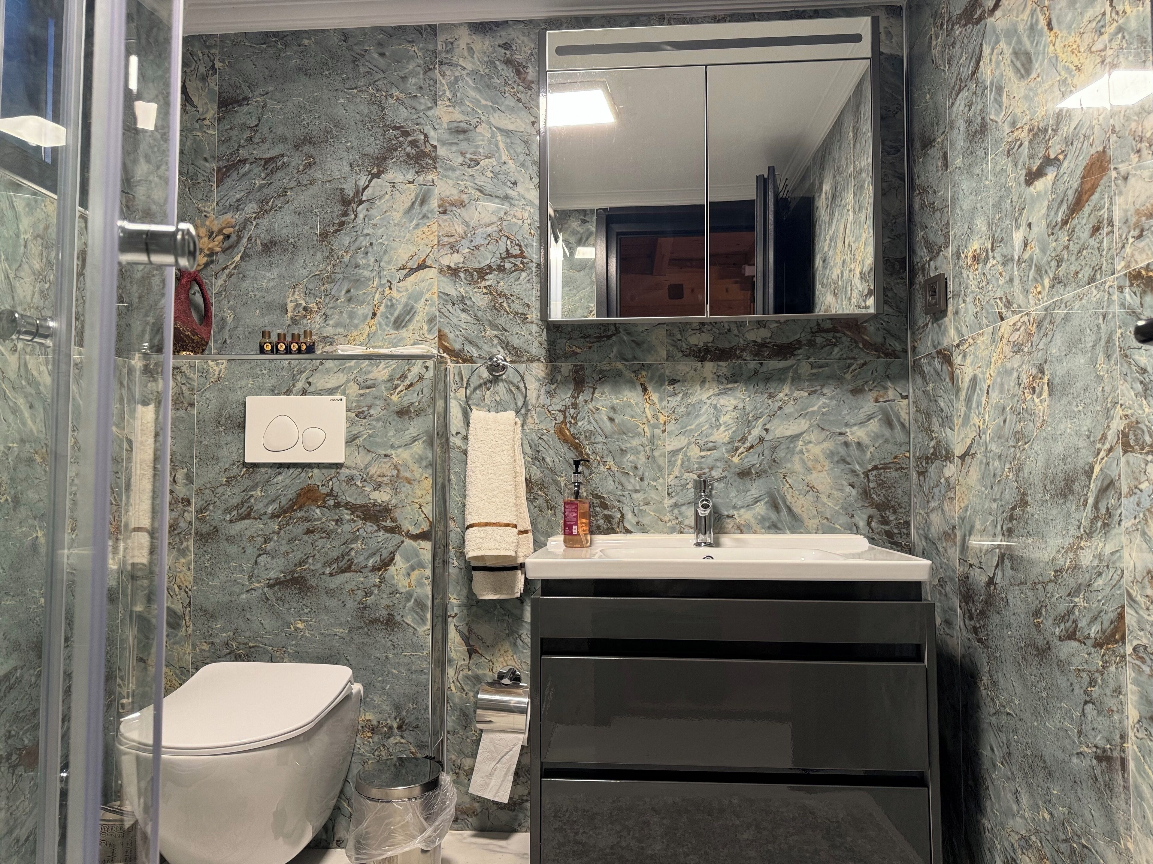 A bathroom with a marble wall, a toilet, a sink with a mirror above it, and a towel rack.