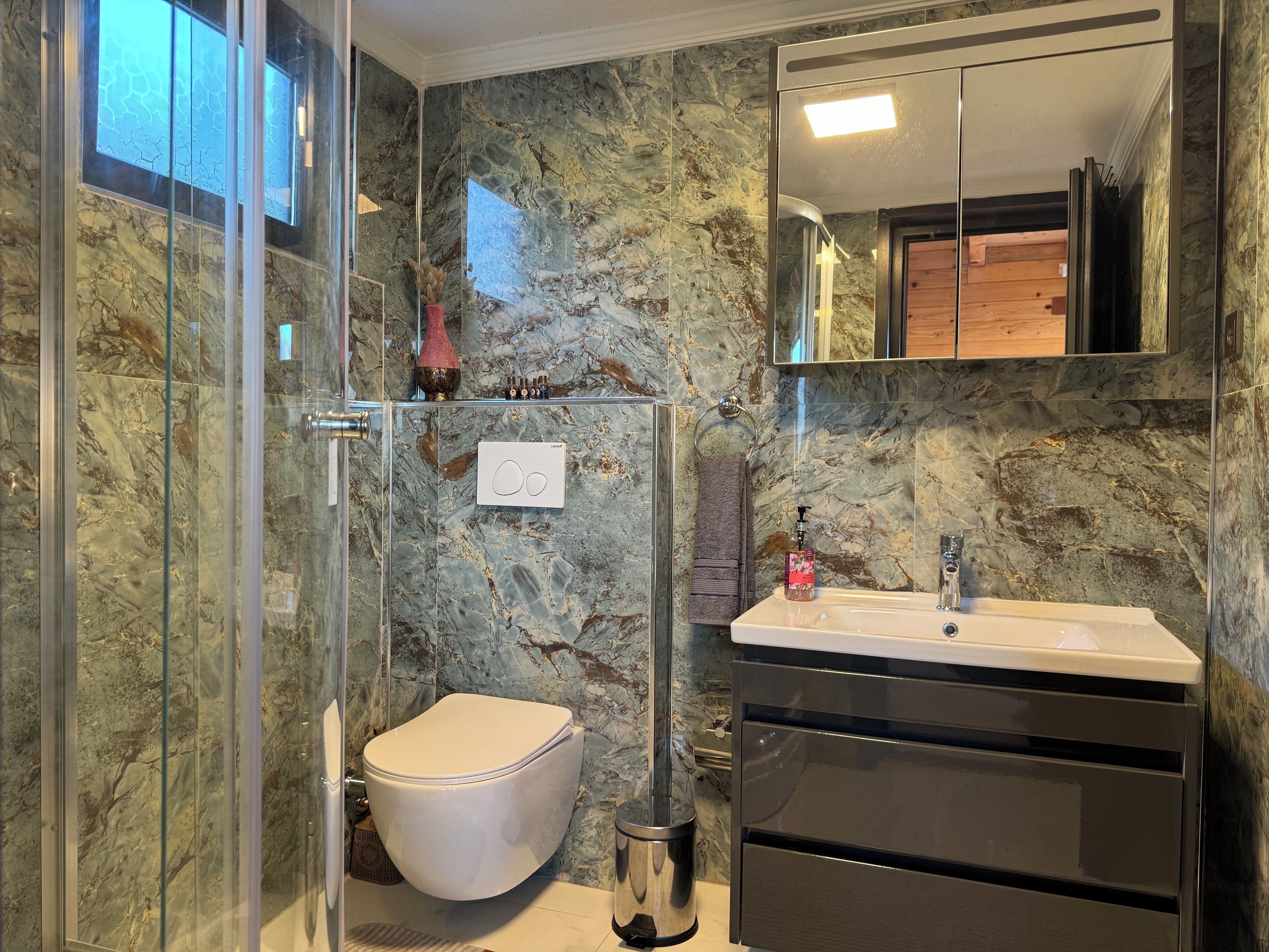 Un baño con cabina de ducha de vidrio, inodoro, lavabo con espejo y paredes de mármol.