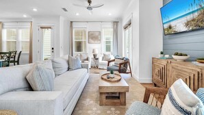A spacious living room with a large flat-screen TV, a ceiling fan, and a view of the beach through the windows.