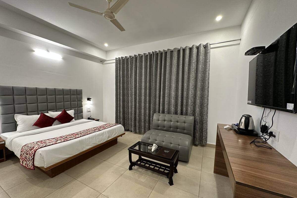 A modern hotel room with a bed, a grey armchair, a small table, a TV, and a ceiling fan.