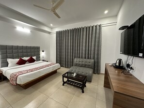 A modern hotel room with a bed, a grey armchair, a small table, a TV, and a ceiling fan. - Gezellig Inn - Magic Mountain (Bhuntar)