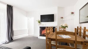 A modern living room with a fireplace, a dining table with chairs, a television mounted on the wall, and a large window with curtains.