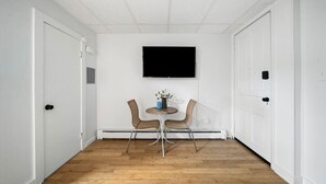 A room with a wooden floor, white walls, a mounted TV, two wicker chairs, and a small table with a vase.