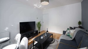Ett modernt vardagsrum med en platt-TV, ett svart soffbord, en grå soffa och en krukväxt.