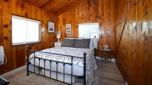 A wooden-paneled bedroom with a bed, two windows, a nightstand, and a small table with a lamp. - Bear Paw- Lake Area  located in Willow Cove (Bass Lake)