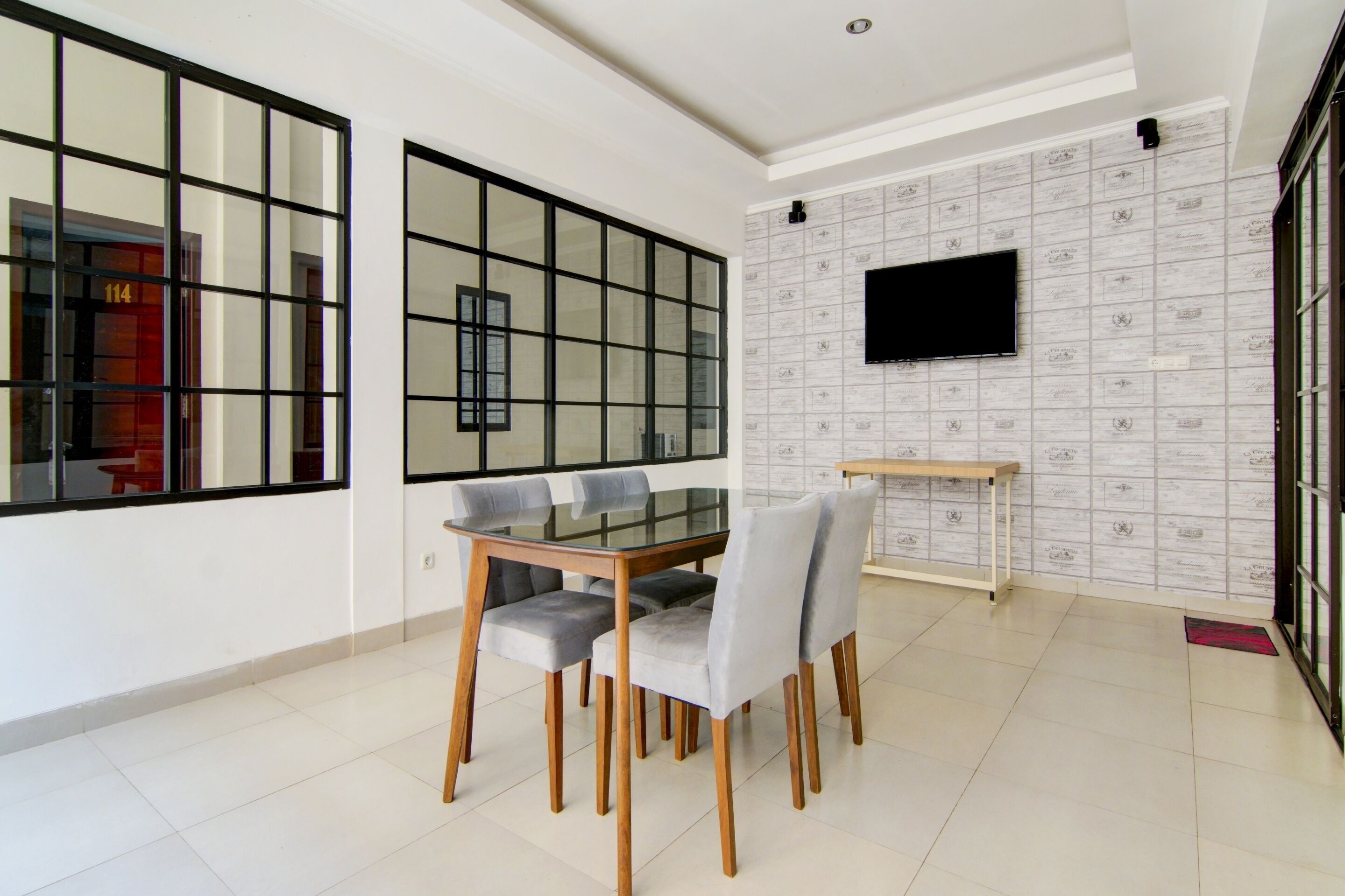 A dining area with a glass-top table and four chairs, a flat-screen TV mounted on a patterned wall, and a bench against the wall.