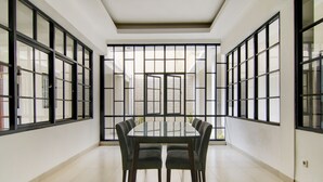 Een ruime kamer met een grote glazen tafel omringd door stoelen, grote ramen met zwarte kaders en een betegelde vloer.