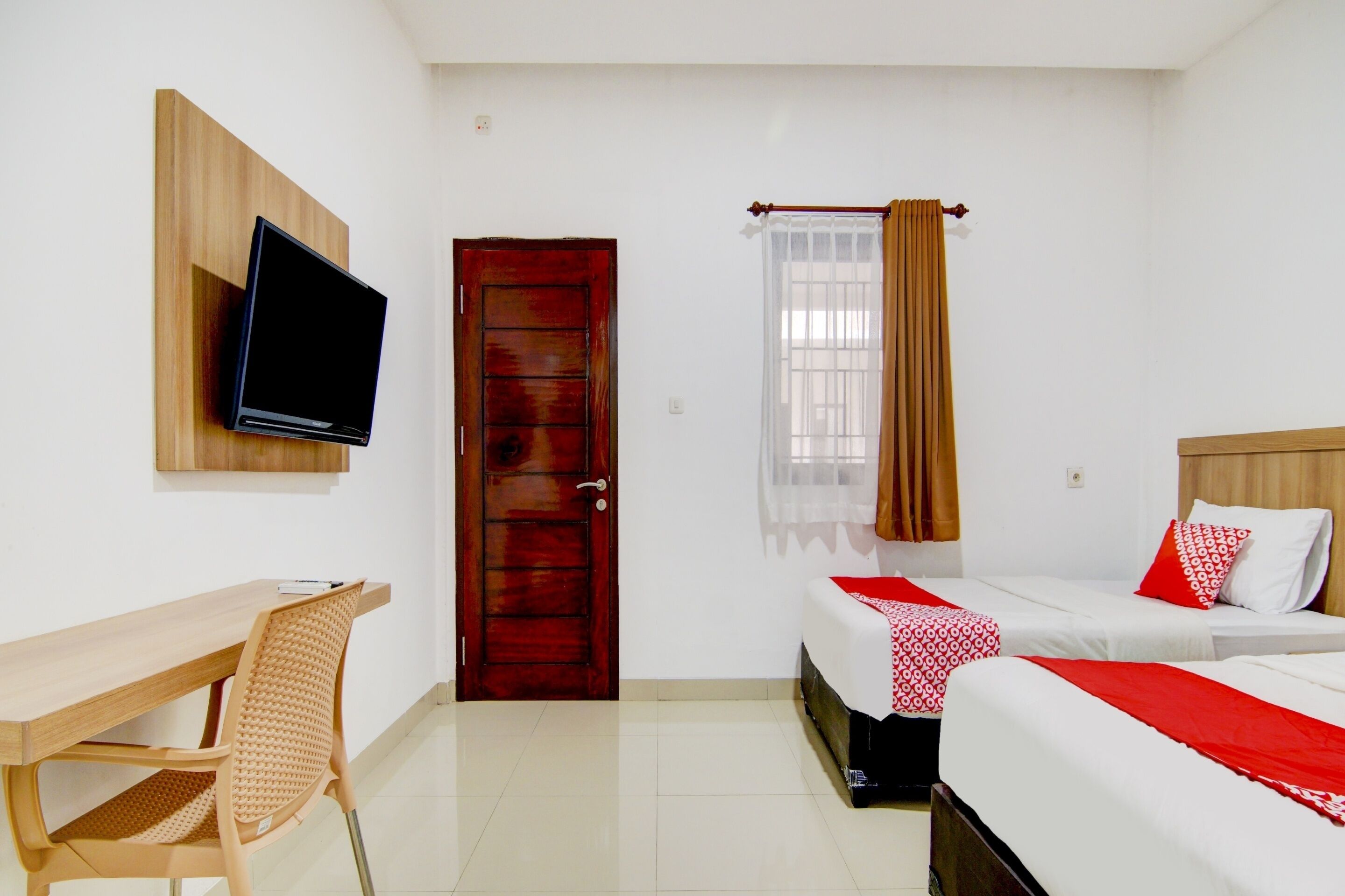 A hotel room with a wooden desk, a television mounted on the wall, two beds with red and white bedding, a wooden door, and a window with curtains.