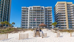 A beachfront hotel with multiple balconies, a flag, and a wooden walkway leading to a gazebo. - Romar Tower Condos (Orange Beach)