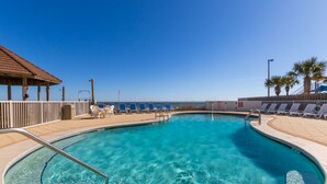 A clear blue pool with a wooden deck and lounge chairs, a covered seating area, and a view of the ocean. - Romar Tower Condos (Orange Beach)