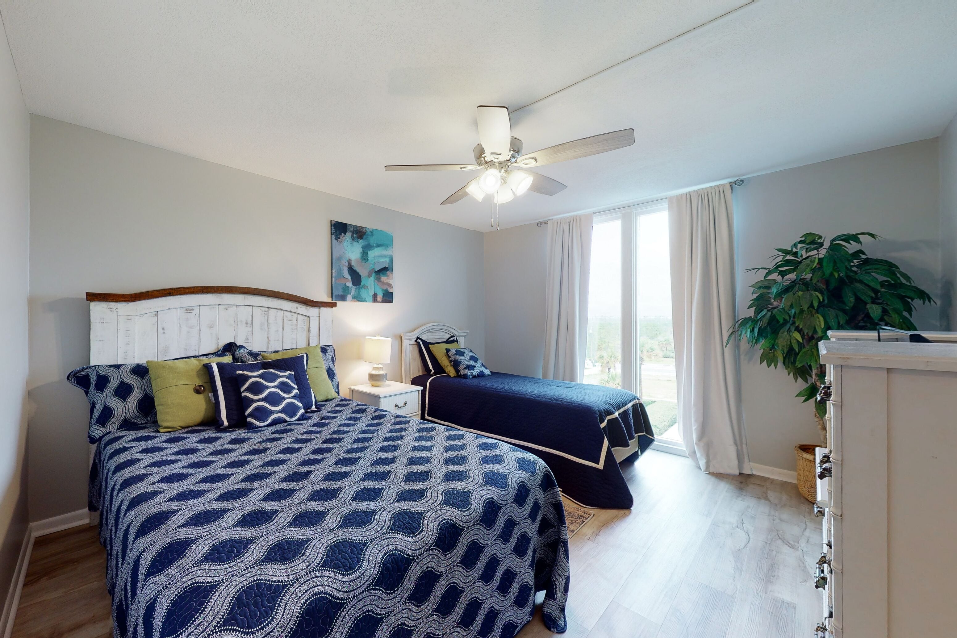 A bedroom with two beds, a ceiling fan, a nightstand, a lamp, a plant, and a window with curtains.