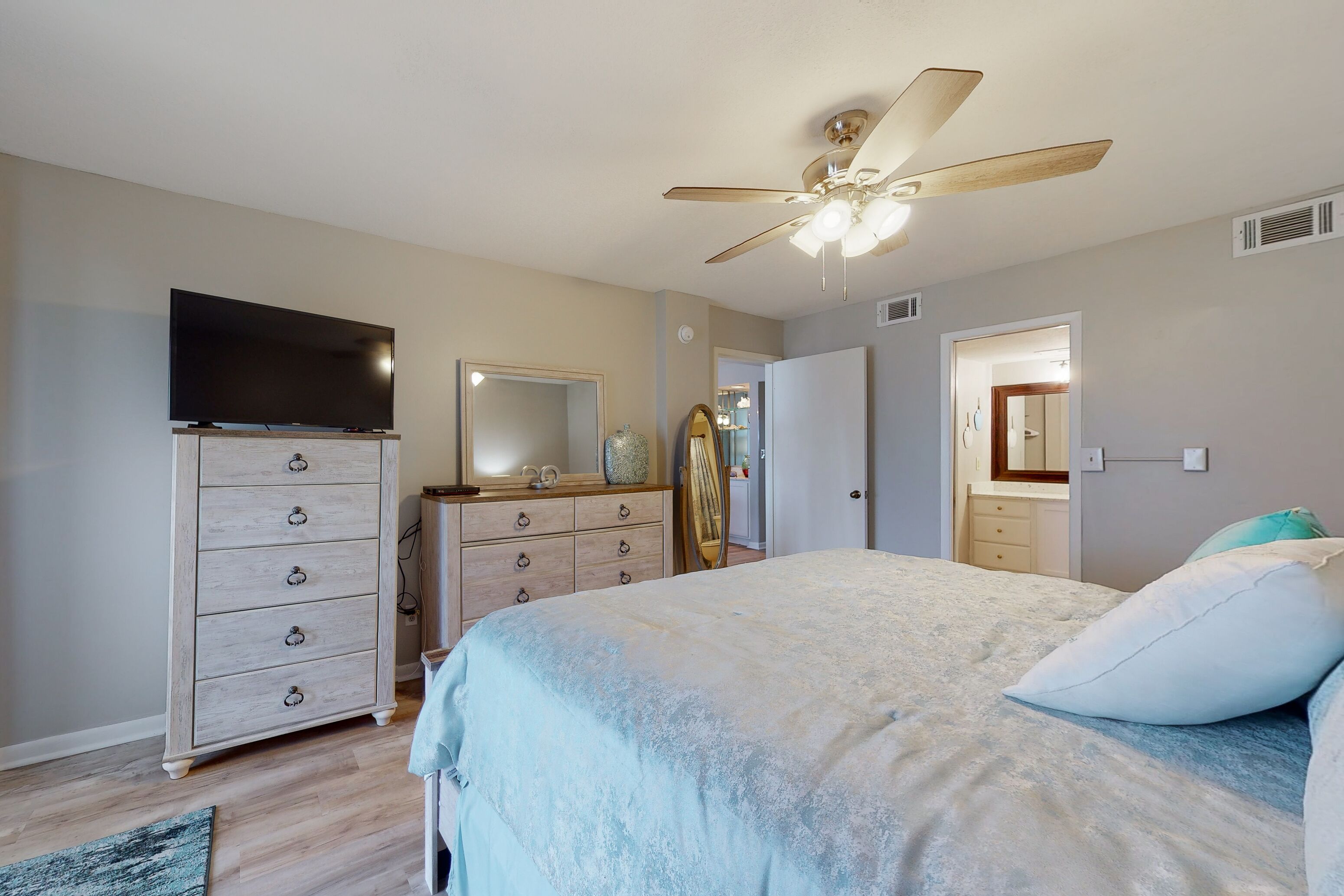 A bedroom with a bed, dresser, TV, ceiling fan, and a door leading to another room.