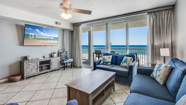 A spacious living room with a large TV, comfortable seating, a ceiling fan, and a view of the ocean. - Romar Tower Condos (Orange Beach)