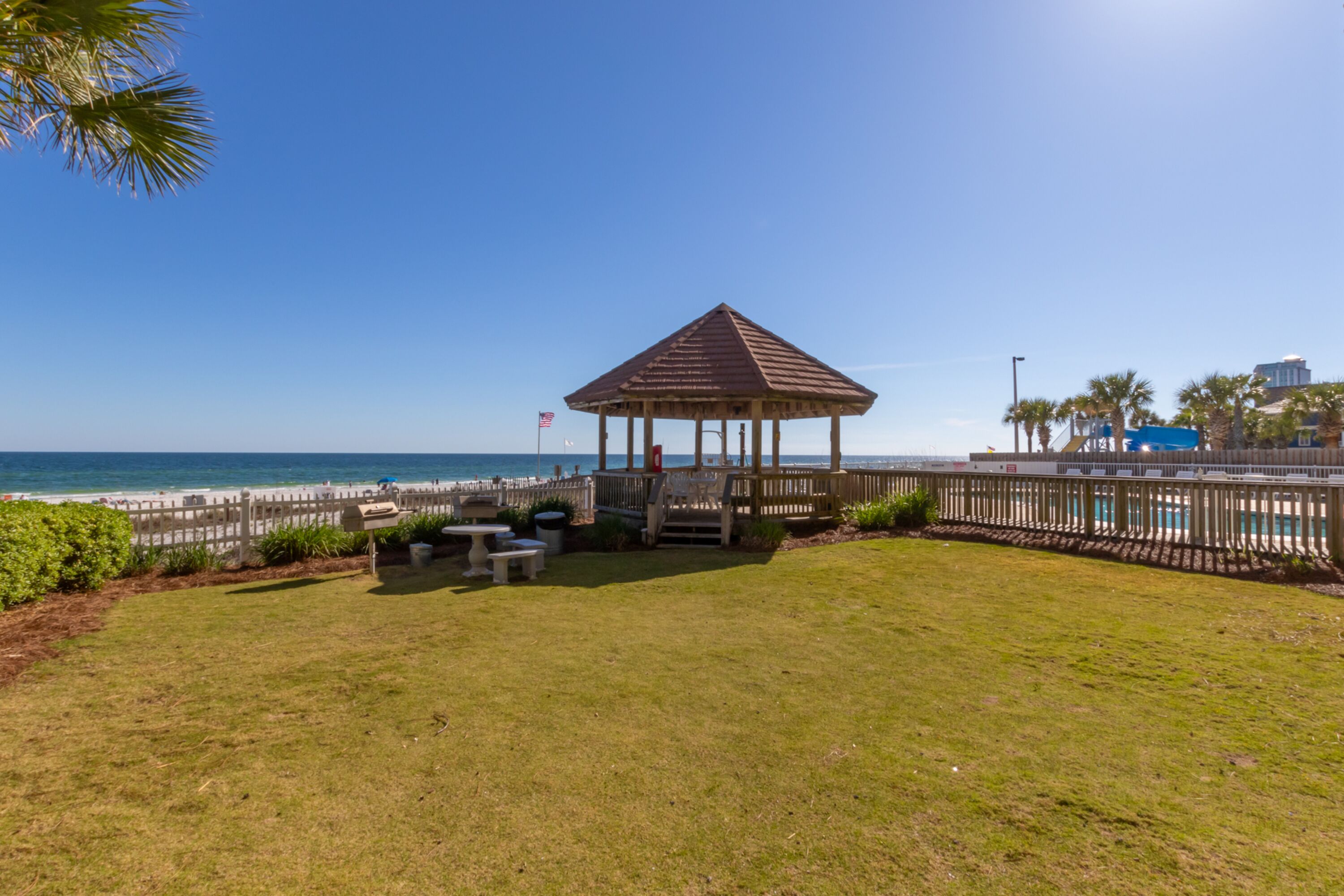 A gazebo with a wooden roof, surrounded by a wooden fence, and a grassy area with a bench and table.