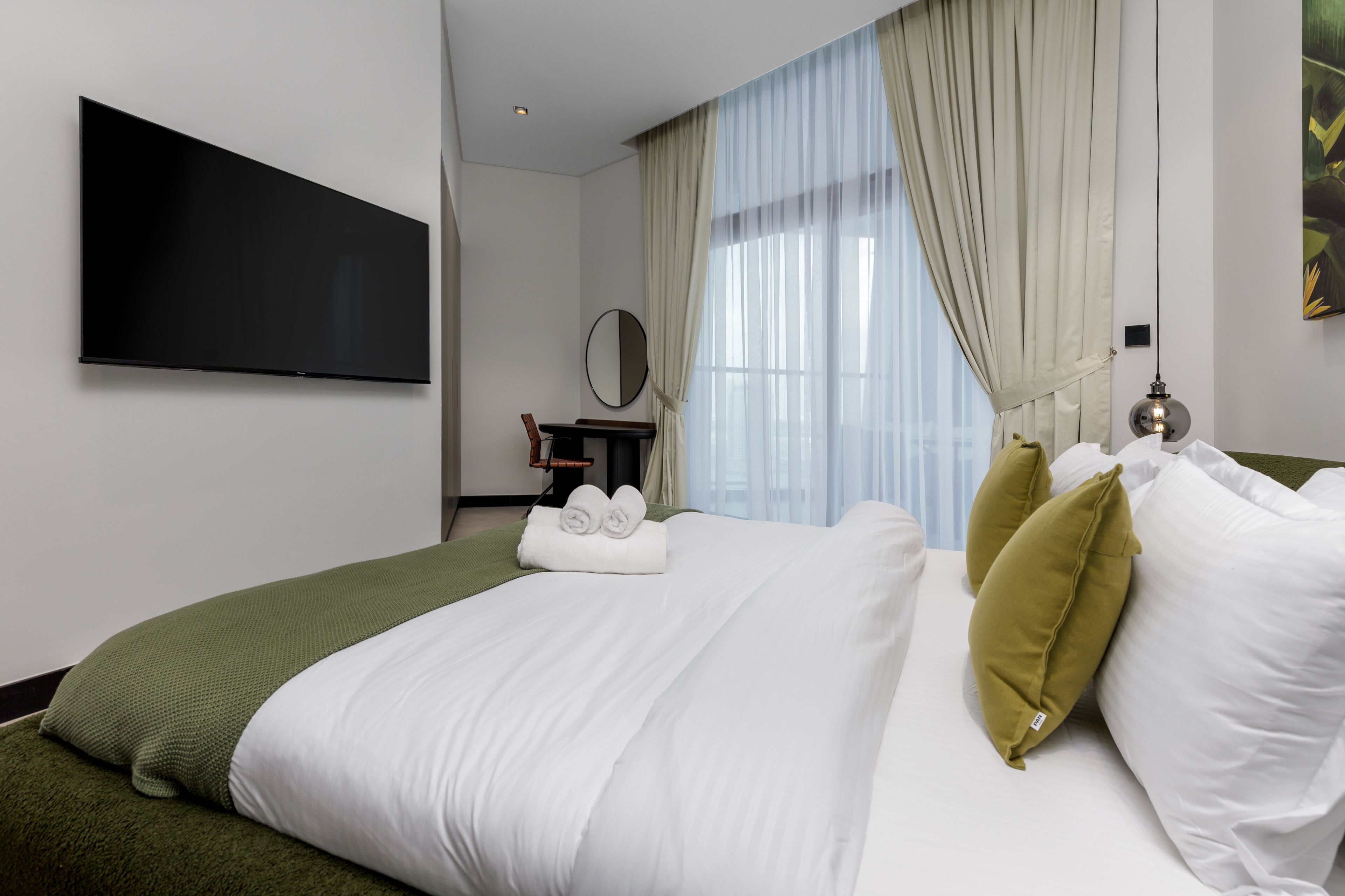 A hotel room with a large bed, a flat-screen TV mounted on the wall, a round mirror, and a window with sheer curtains.