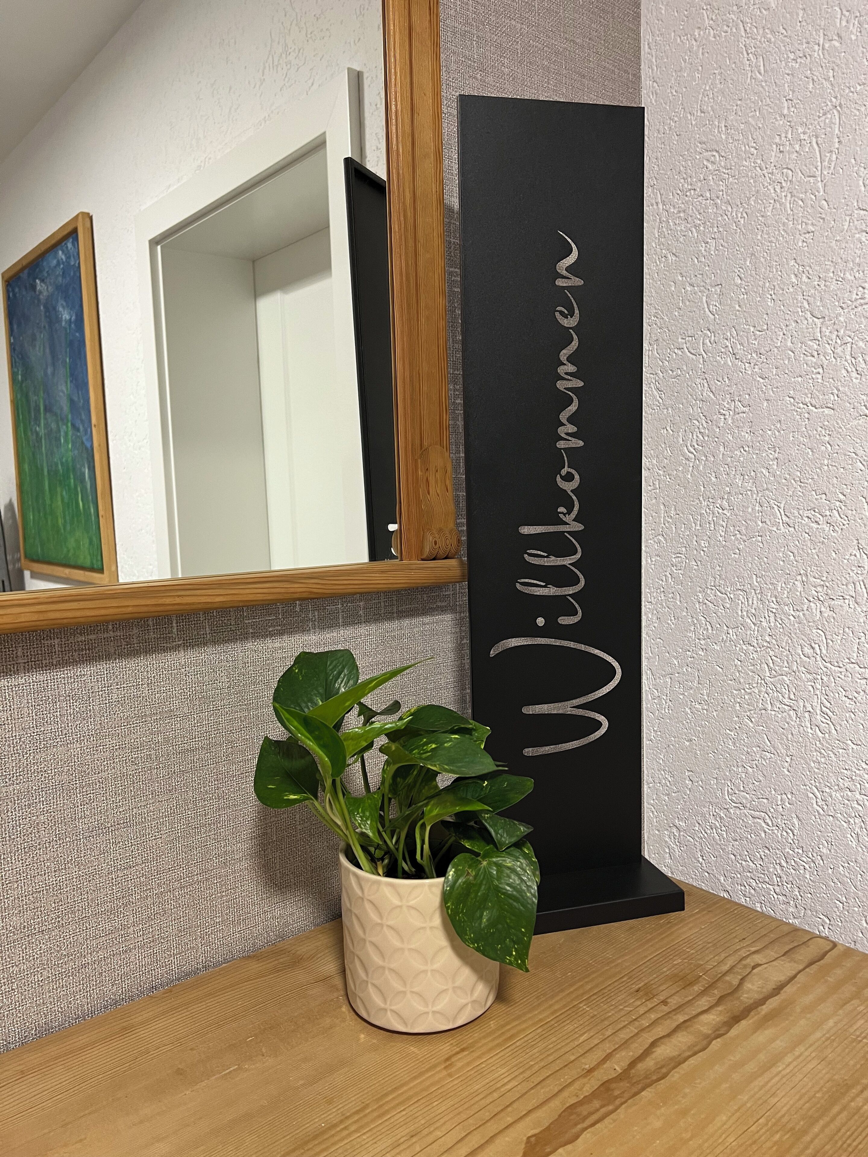 En krukväxt står på ett träbord, och bredvid den finns en svart skylt med texten ”Willkommen” i vita bokstäver.