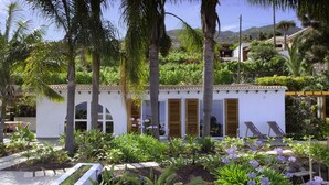 Un pequeño edificio blanco con ventanas arqueadas y contraventanas de madera, rodeado de exuberante vegetación y plantas en flor.