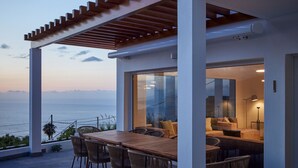 Un espacio exterior moderno para comer, con un pérgola de madera, una mesa grande y sillas, con vistas a un paisaje marino.