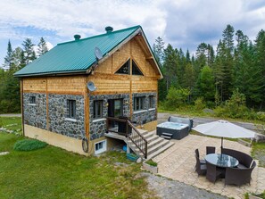 Una cabaña con fachada de piedra, techo verde y un patio con mobiliario exterior.