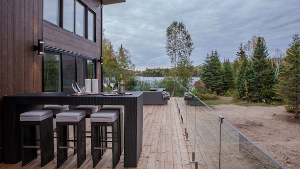 A modern outdoor deck with a wooden floor, black wicker furniture, and a view of a lake and forest. - Magnificent chalet with a contemporary design: dream location for an unforgettable holiday (Notre-Dame-de-la-Merci)