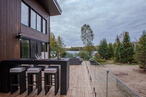 Une terrasse extérieure moderne avec un plancher en bois, des meubles en osier noir et une vue sur un lac et une forêt.