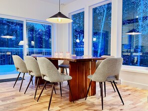 Comedor con mesa de madera y cuatro sillas, ventanas amplias con vista a árboles y una lámpara colgante.